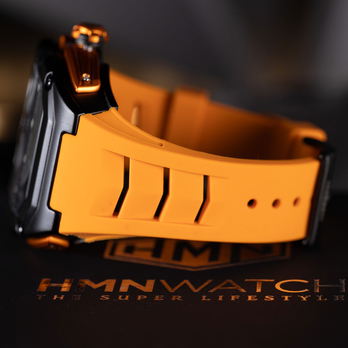 Close-up of a watch with an orange strap on a dark background, featuring the brand 'HMNWATCH'.