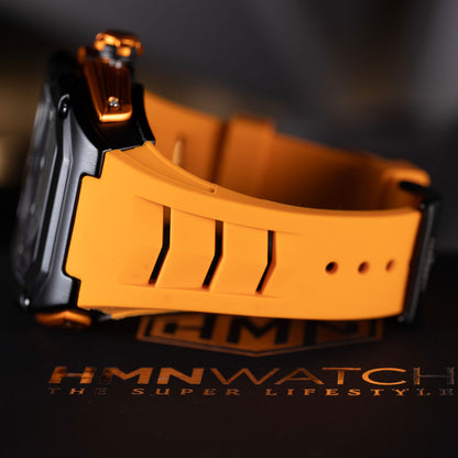 Close-up of a watch with an orange strap on a dark background, featuring the brand 'HMNWATCH'.